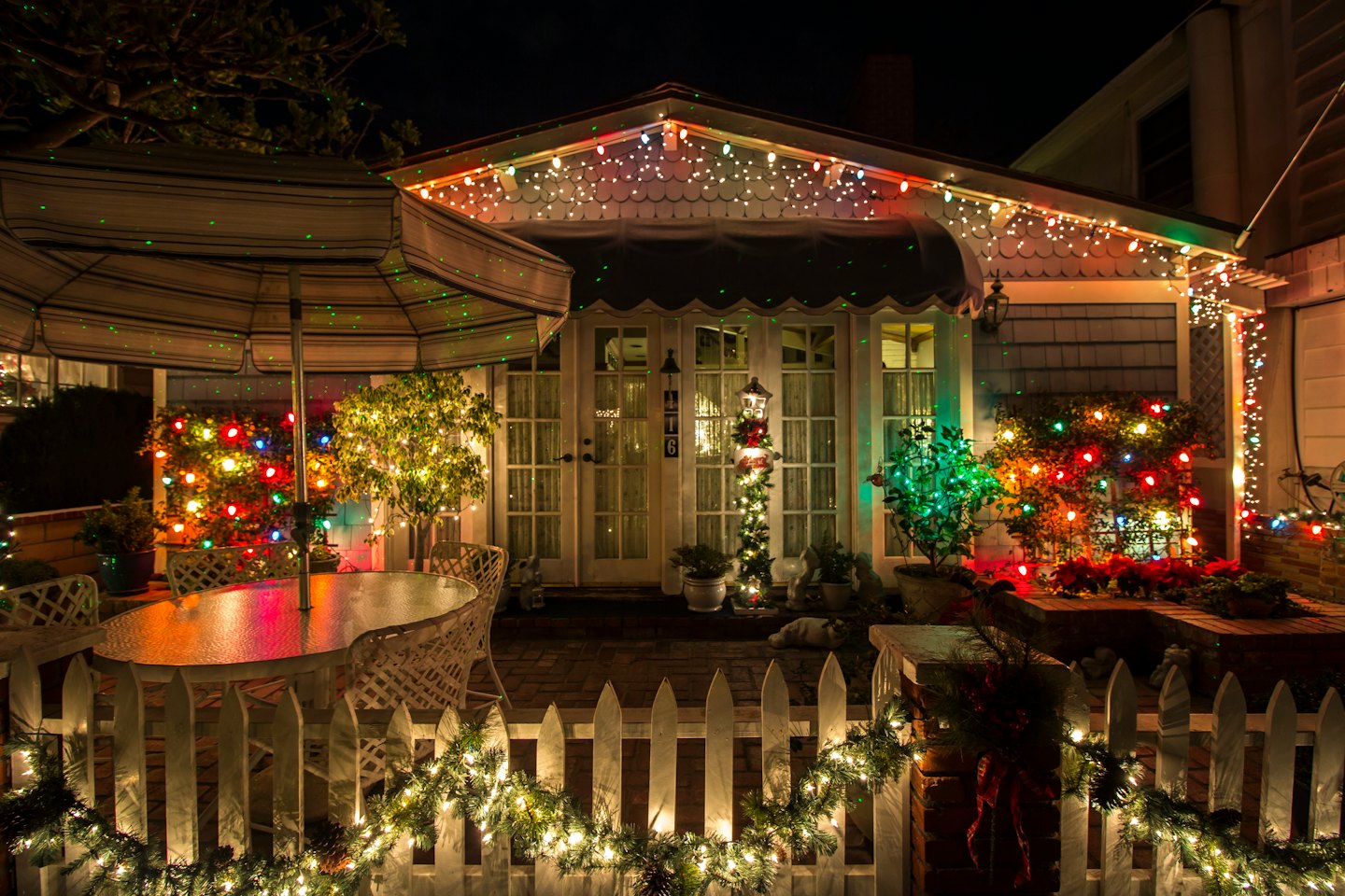 Holiday lights illuminate a house patio and white picket fence.